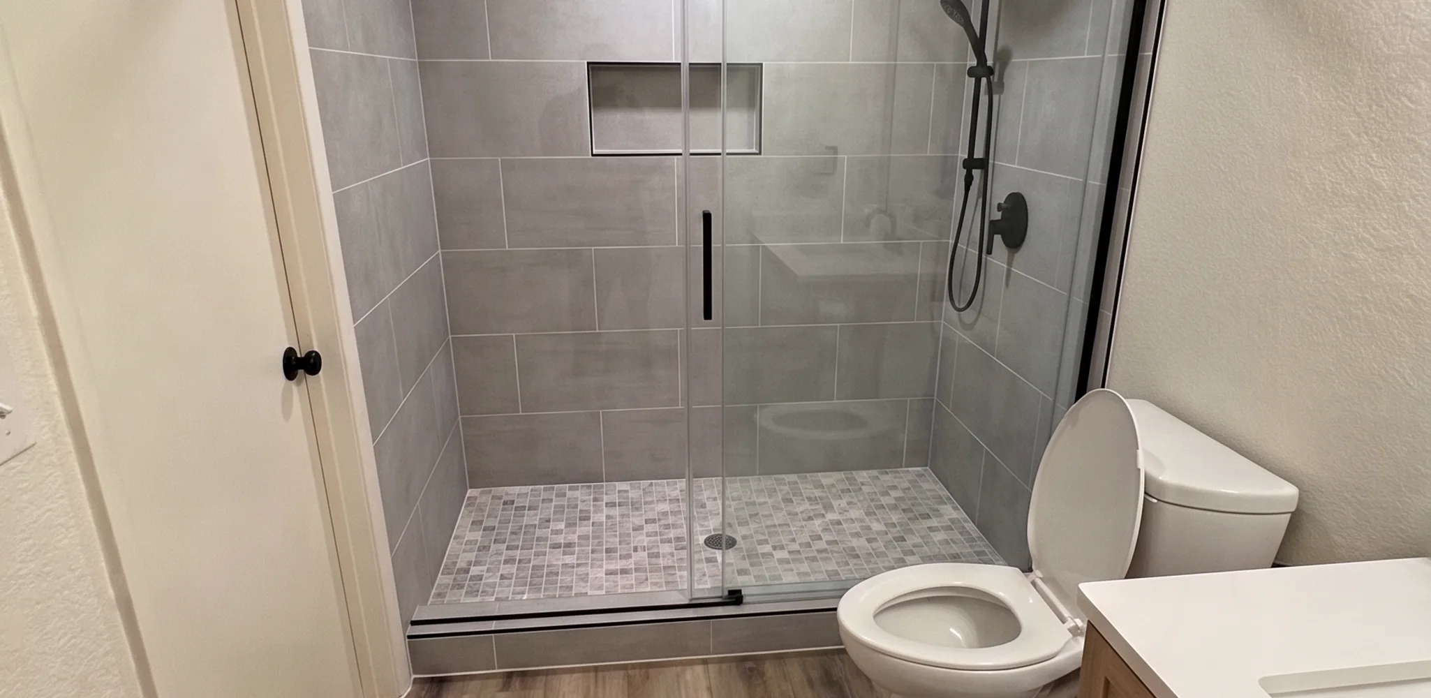 Walk-in shower built in the footprint of a former bathtub alcove, with large format porcelain tile, a mosaic tile shower floor, a low tiled curb and a two-panel sliding glass door