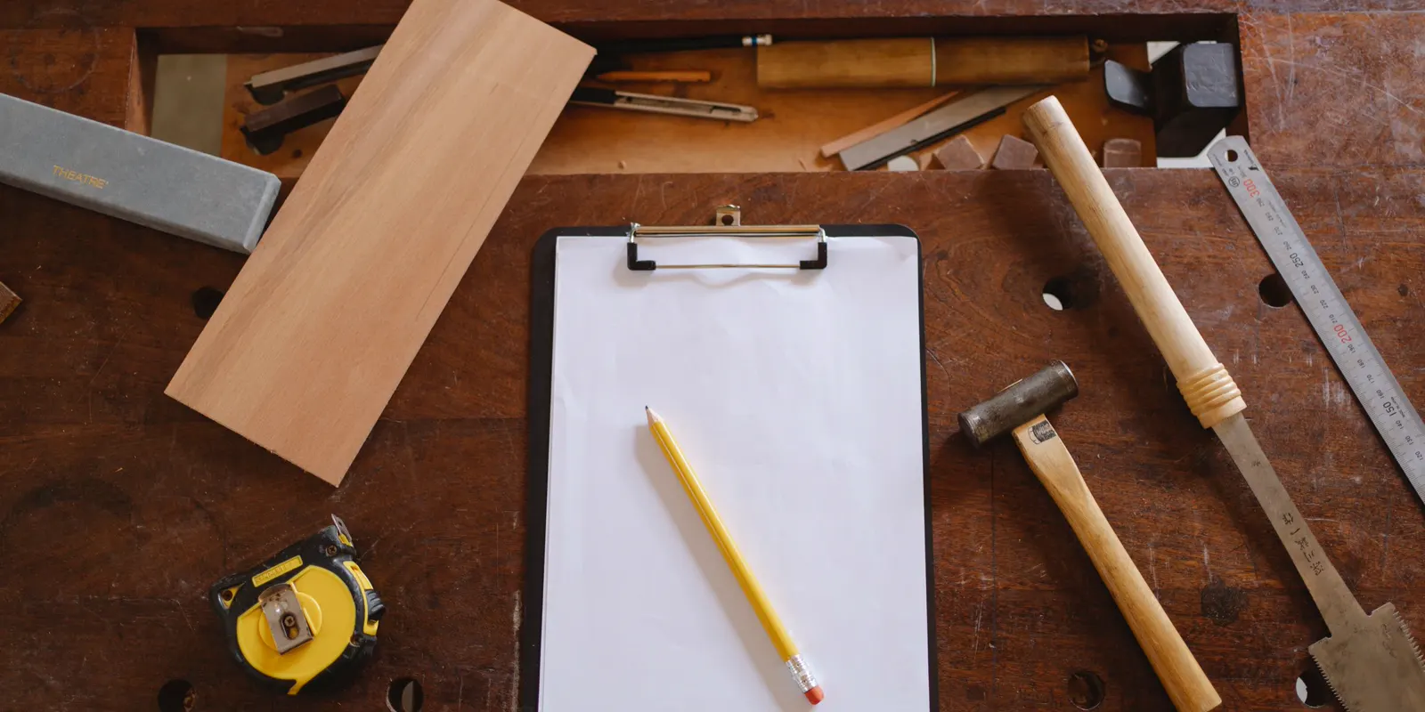 A clipboard, a pencil, a tape measure and hand tools laid out on a wooden bench