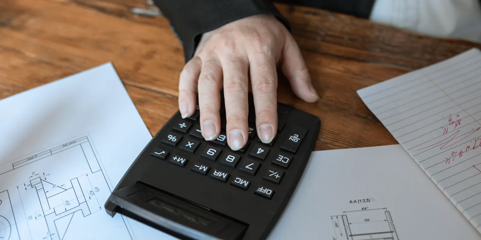 A hand pressing the keys of a calculator that sits on a set of blueprints