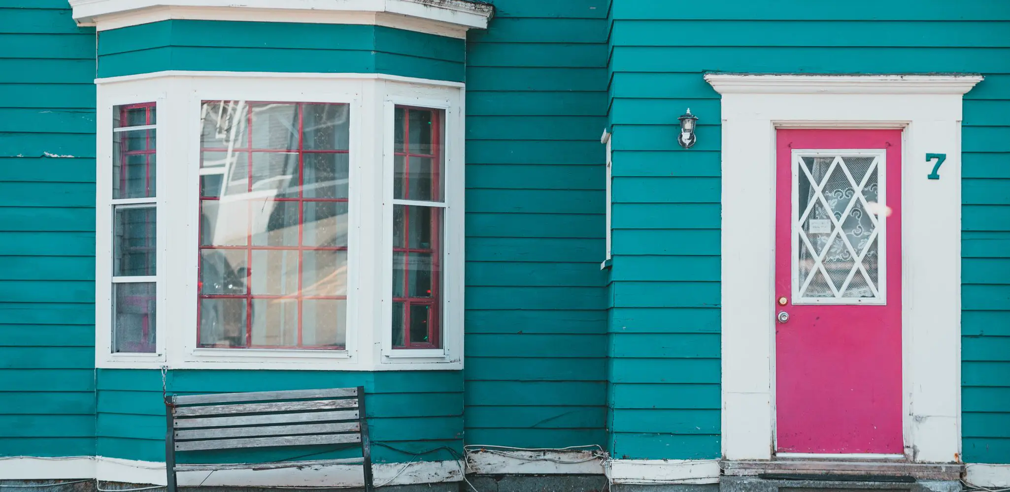 A teal house with a red door, a window and a bench, exterior and front of house