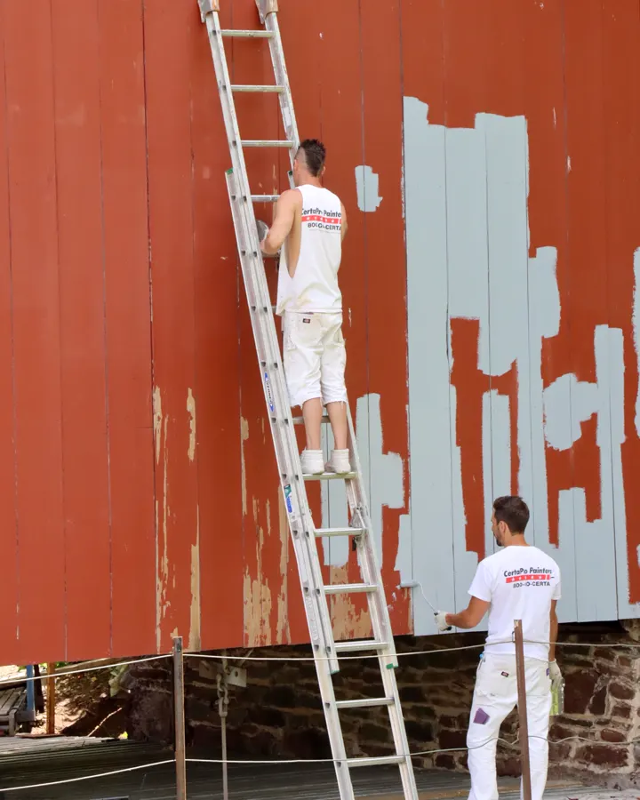 Painters scraping and priming a peeling red wall from a ladder before repainting, exterior painting prep