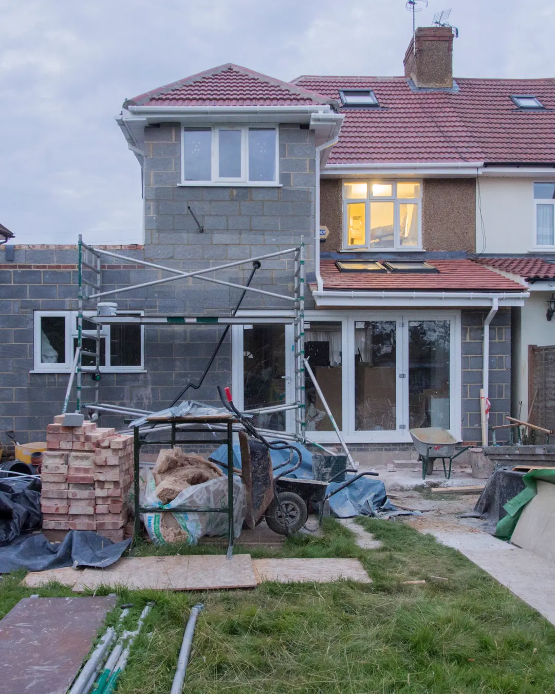 A house with a rear extension under construction, scaffolding and building materials in the yard, room addition