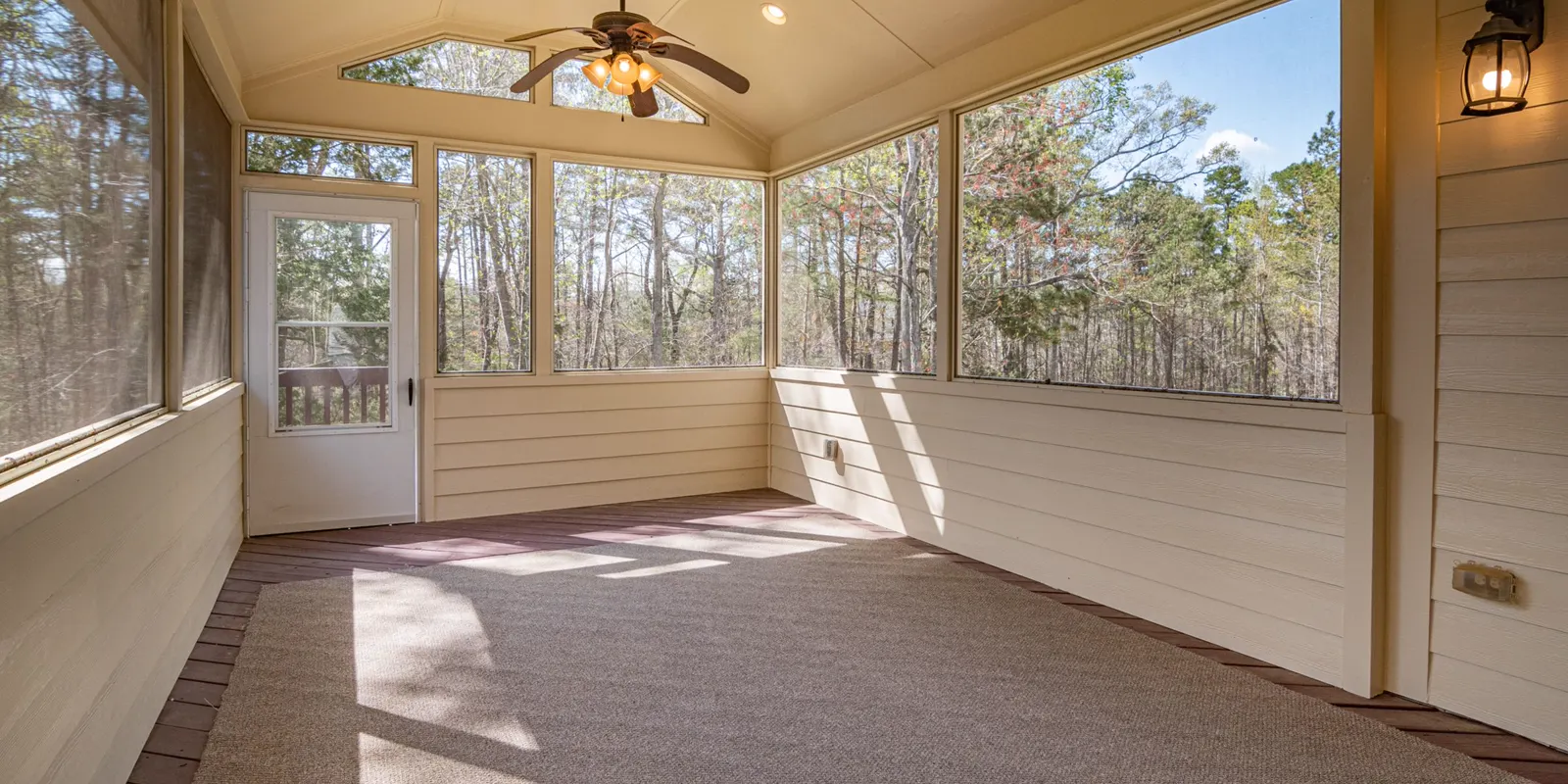 An empty porch with a ceiling fan and glass walls looking into the trees, sunroom and Florida room